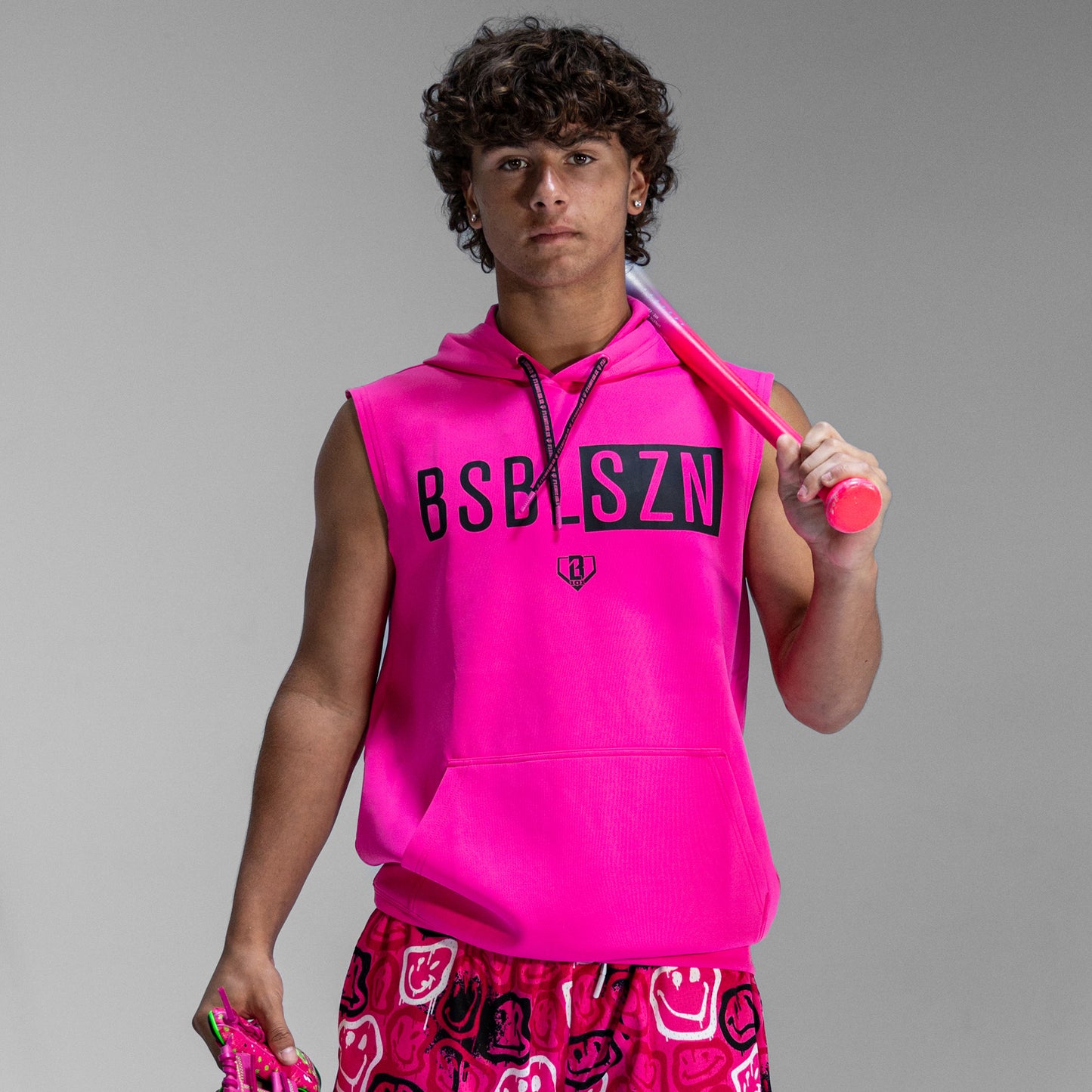 Person wearing a bright pink sleeveless hoodie with 'BSBLSZN' logo, holding a pink bat against a gray background.