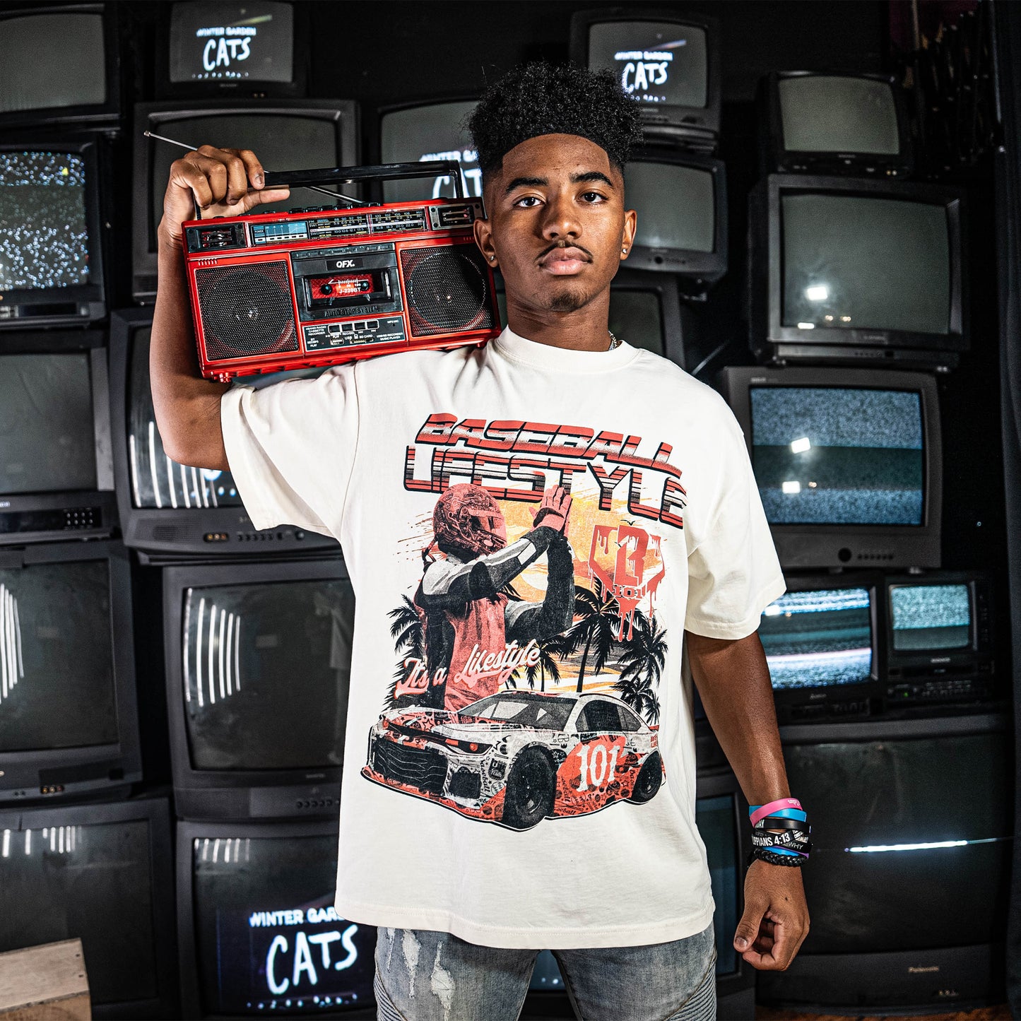 Person wearing a white t-shirt with a graphic design of a car and 'Baseball Lifestyle' text, holding a red boom box standing in a room with stacked tv's.