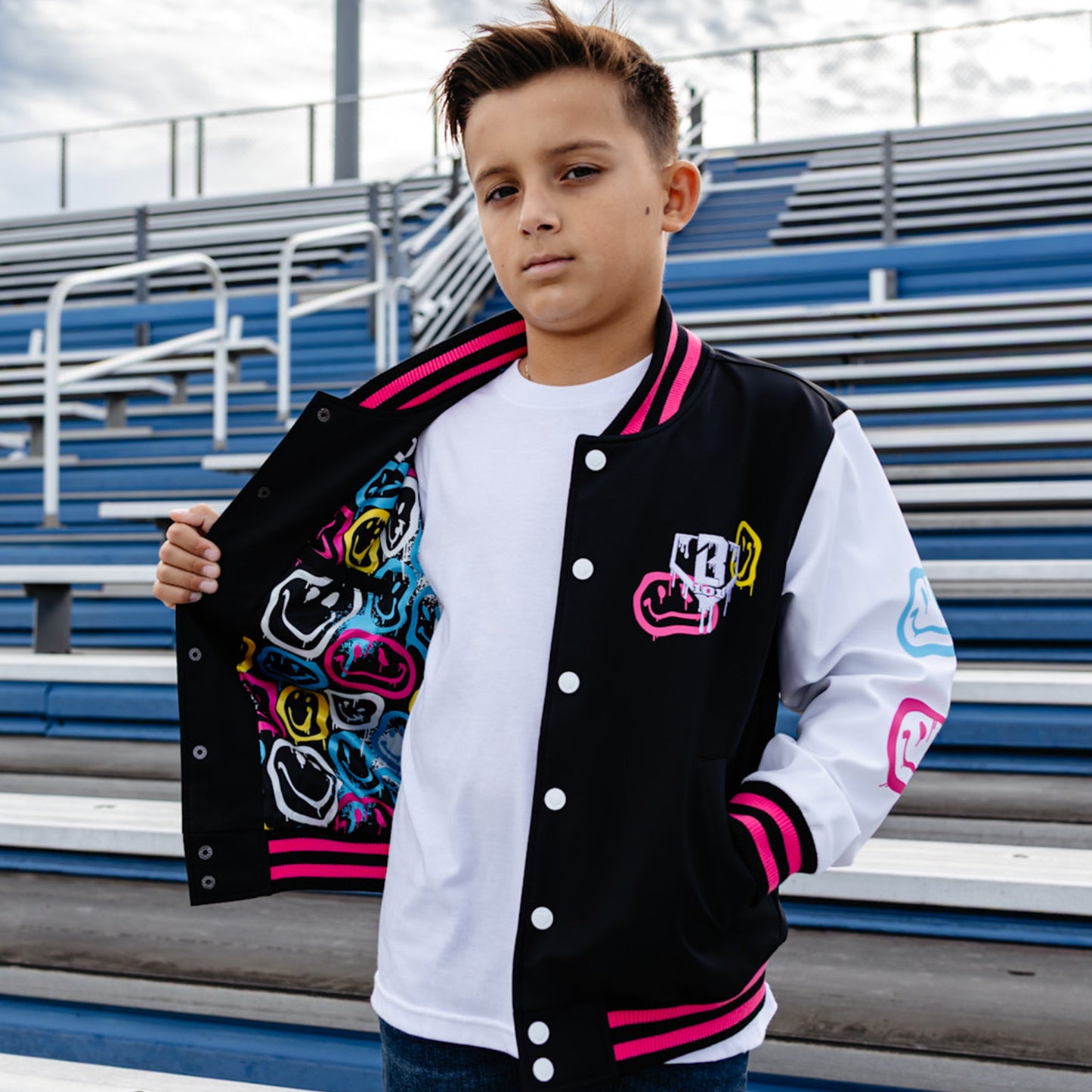 Child wearing a black, pink and white varsity jacket with colorful designs, standing in front of bleachers.