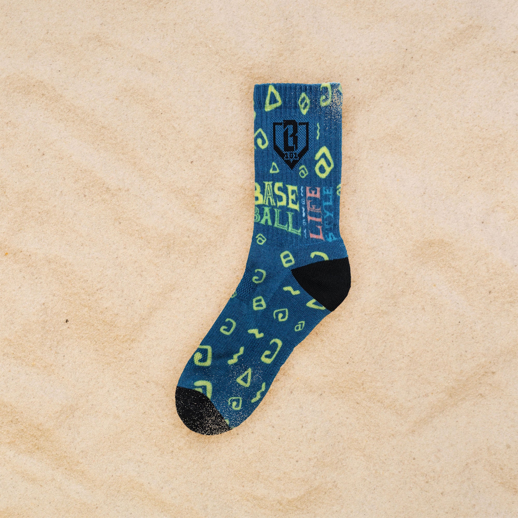 A single blue crew sock with green geometric patterns and text reading 'BASEBALL LIFESTYLE' lies on a sandy beige background.