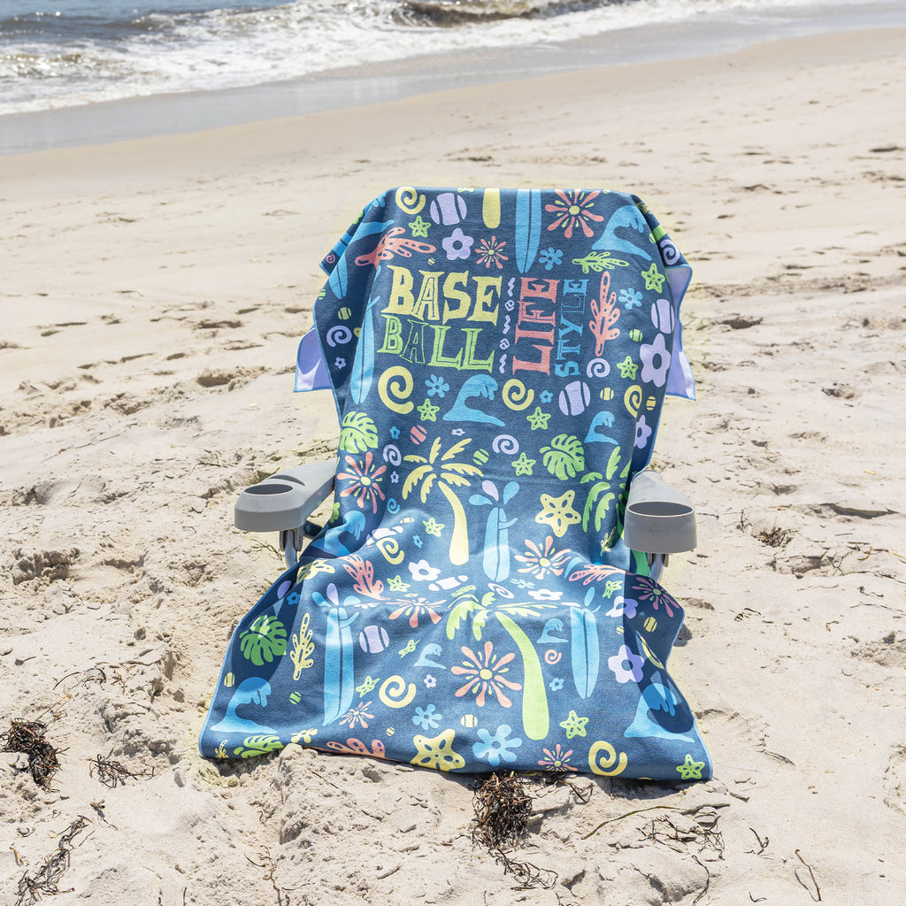 A beach chair with a colorful 'BASEBALL LIFESTYLE' print sits on a sandy beach with the ocean waves in the background.