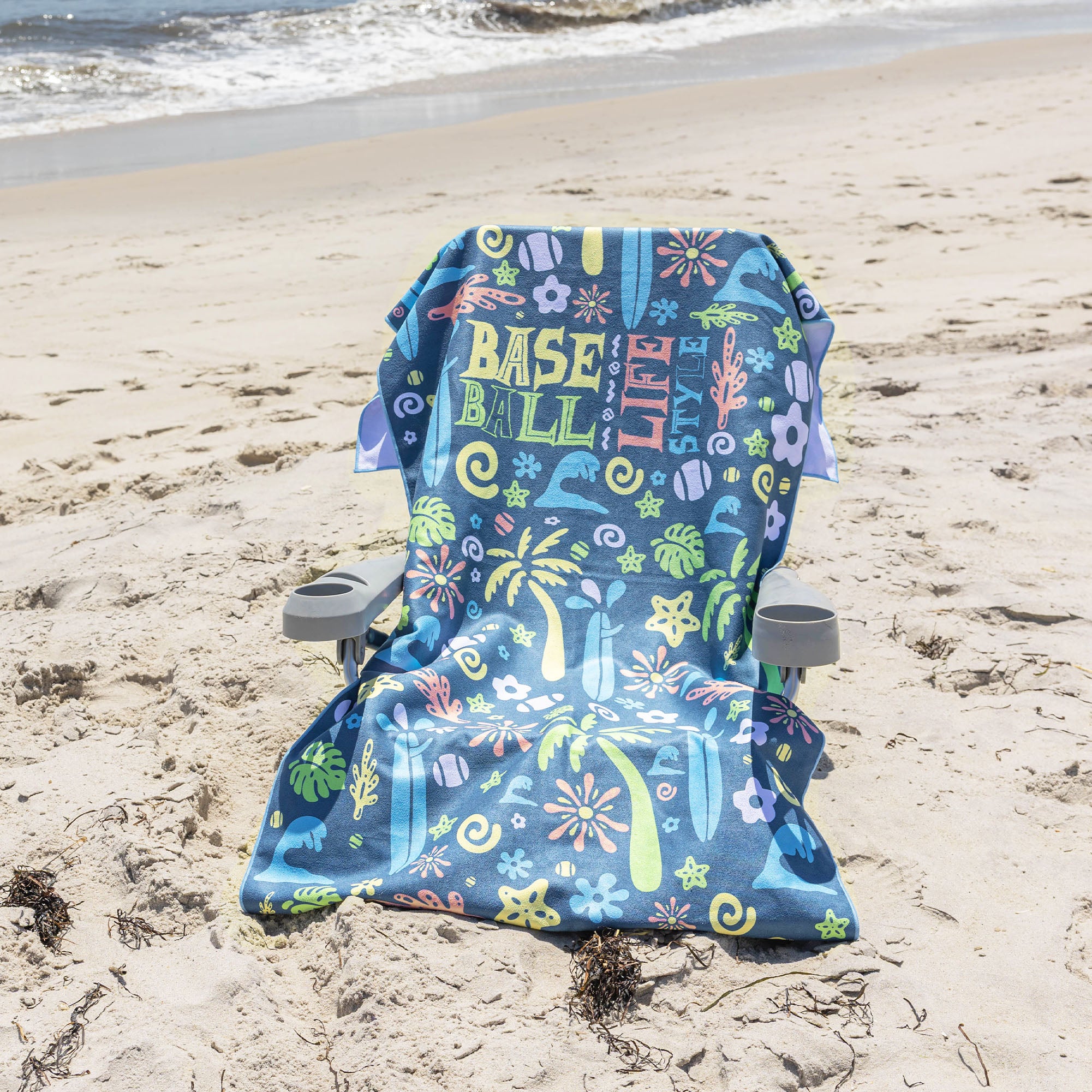 A beach chair with a colorful 'BASEBALL LIFESTYLE' print sits on a sandy beach with the ocean waves in the background.