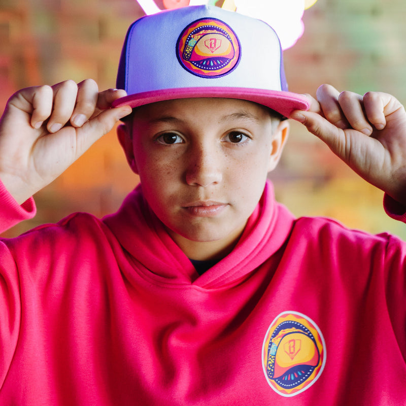 Person wearing a pink and purple cap with  taco graphic design and a matching hoodie, against a blurred outdoor background.