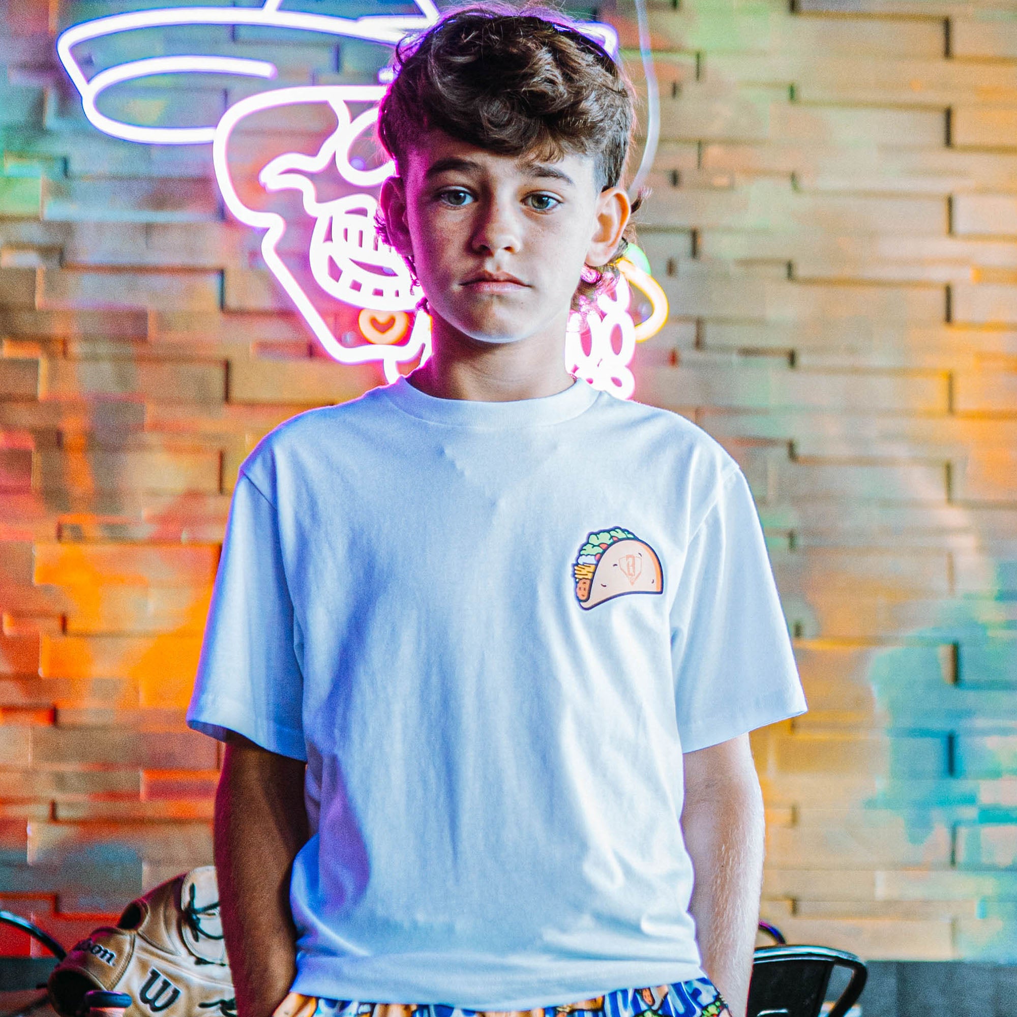 Person wearing a white t-shirt with a taco graphic in front of a colorful neon sign.