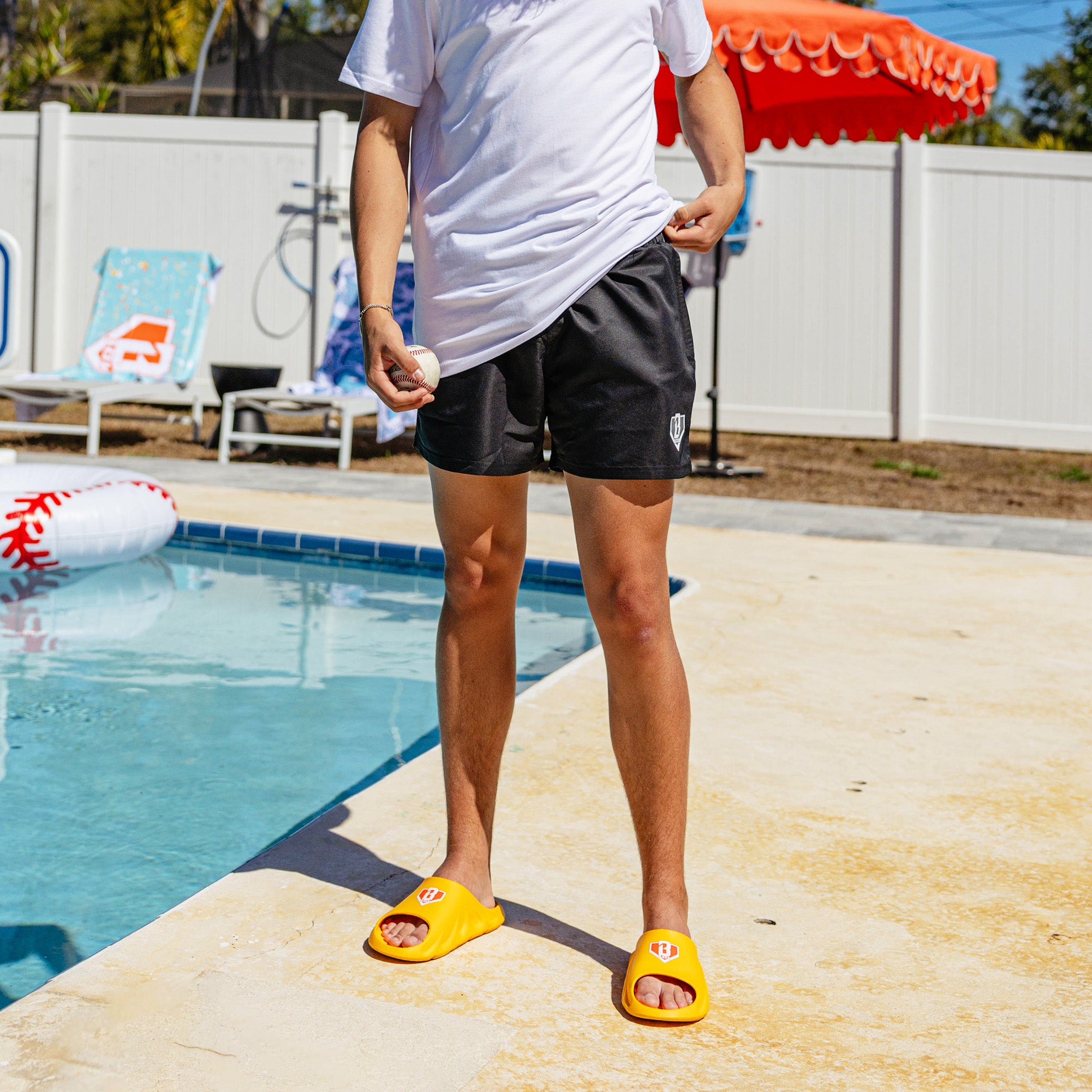 Person wearing BL101 yellow slides and black swim trunks poolside, holding a baseball.
