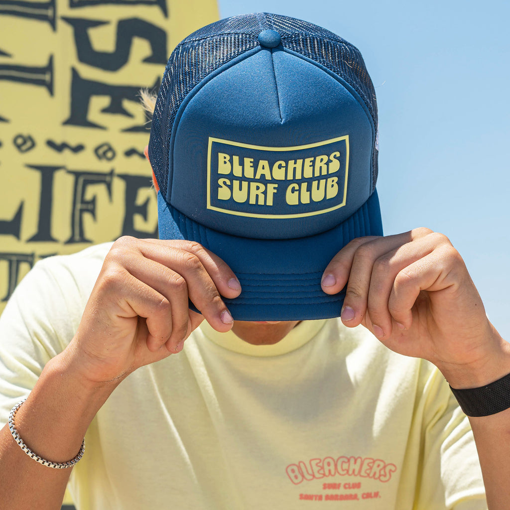 Adult model wearing blue hat with "Bleachers Surf Club" design