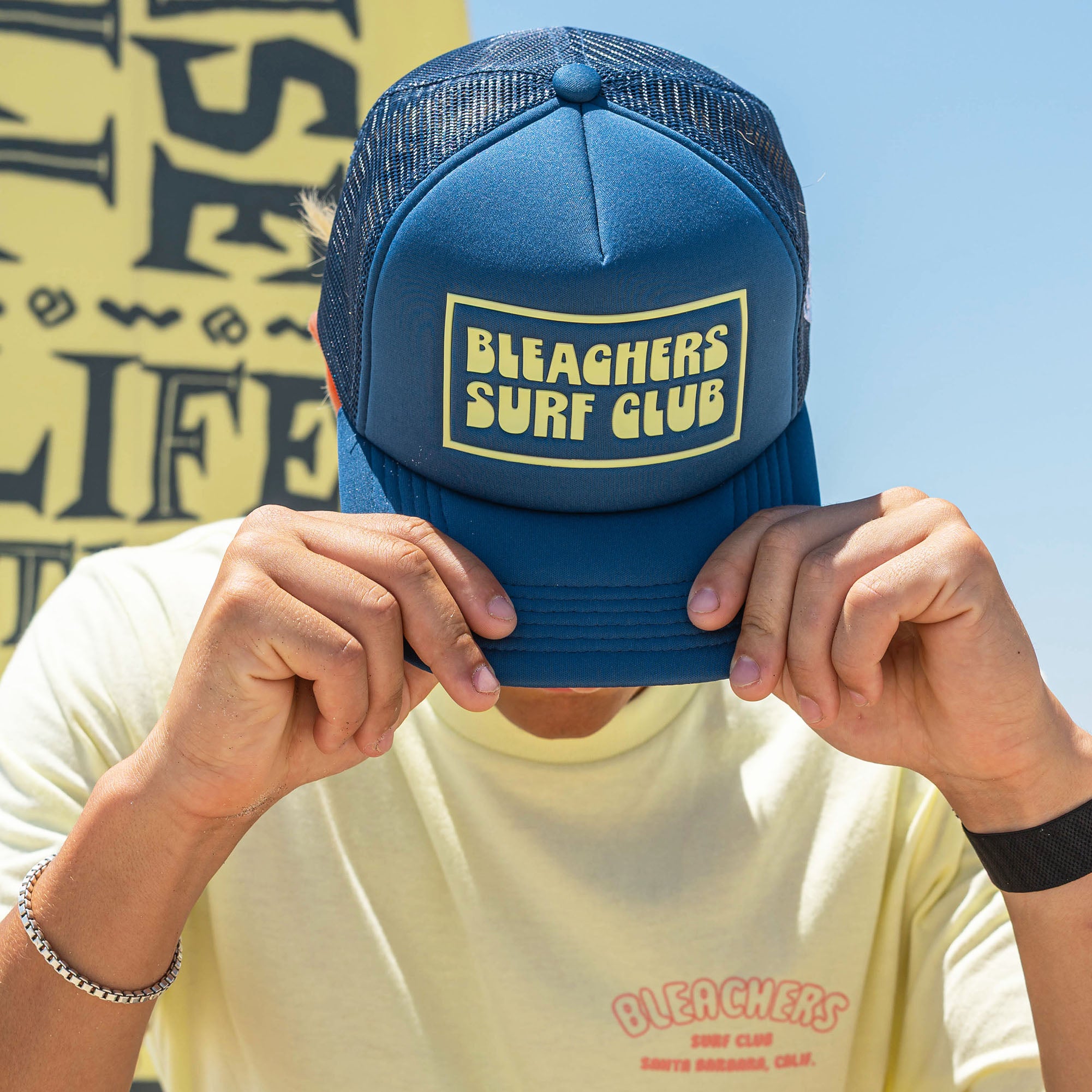 Adult model wearing blue hat with "Bleachers Surf Club" design