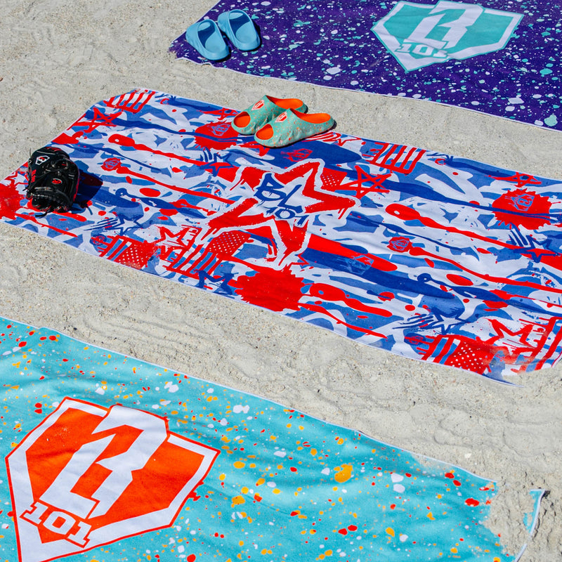 Three BL101 beach towels laid out on sand — red/white/blue graffiti print, purple splatter, and teal splatter — with slides, a baseball glove, and sandals.