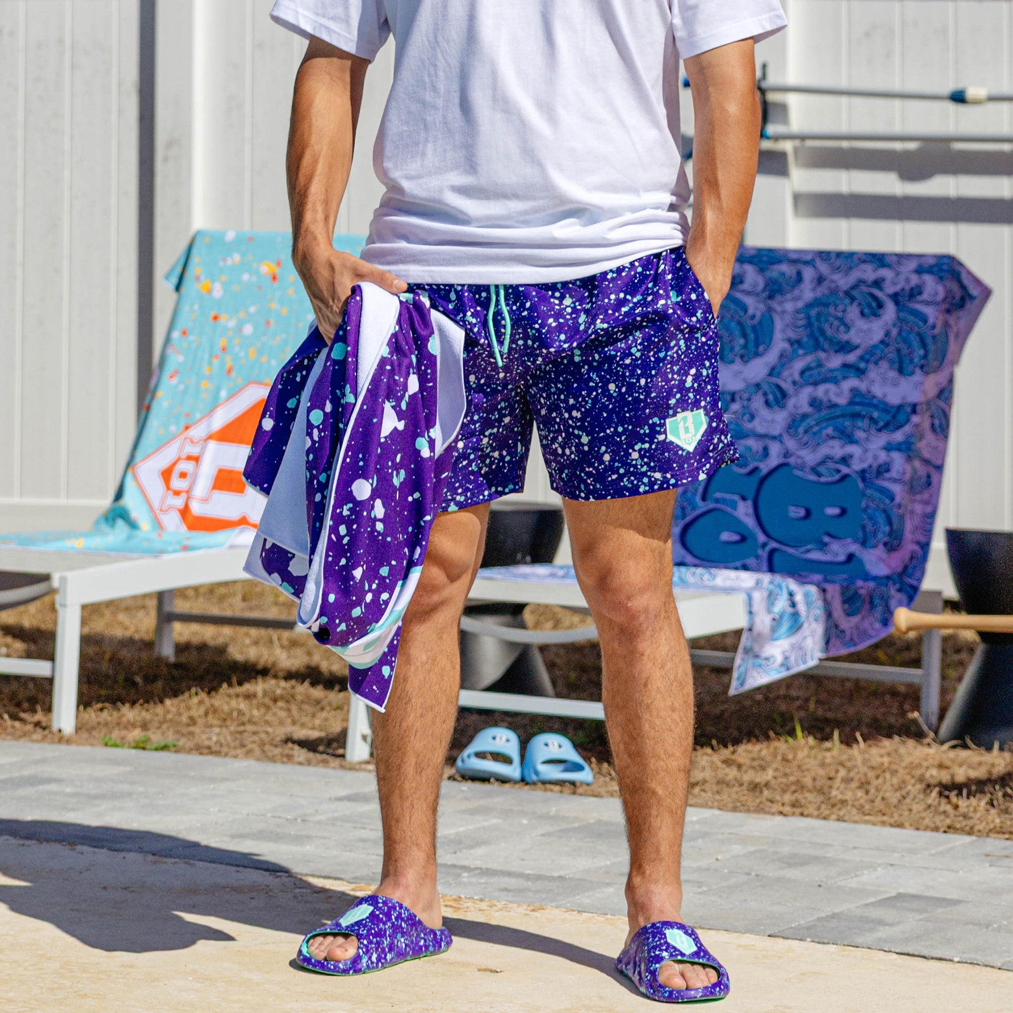 Person wearing BL101 purple splatter slides and matching swim trunks poolside, holding a matching purple splatter towel.
