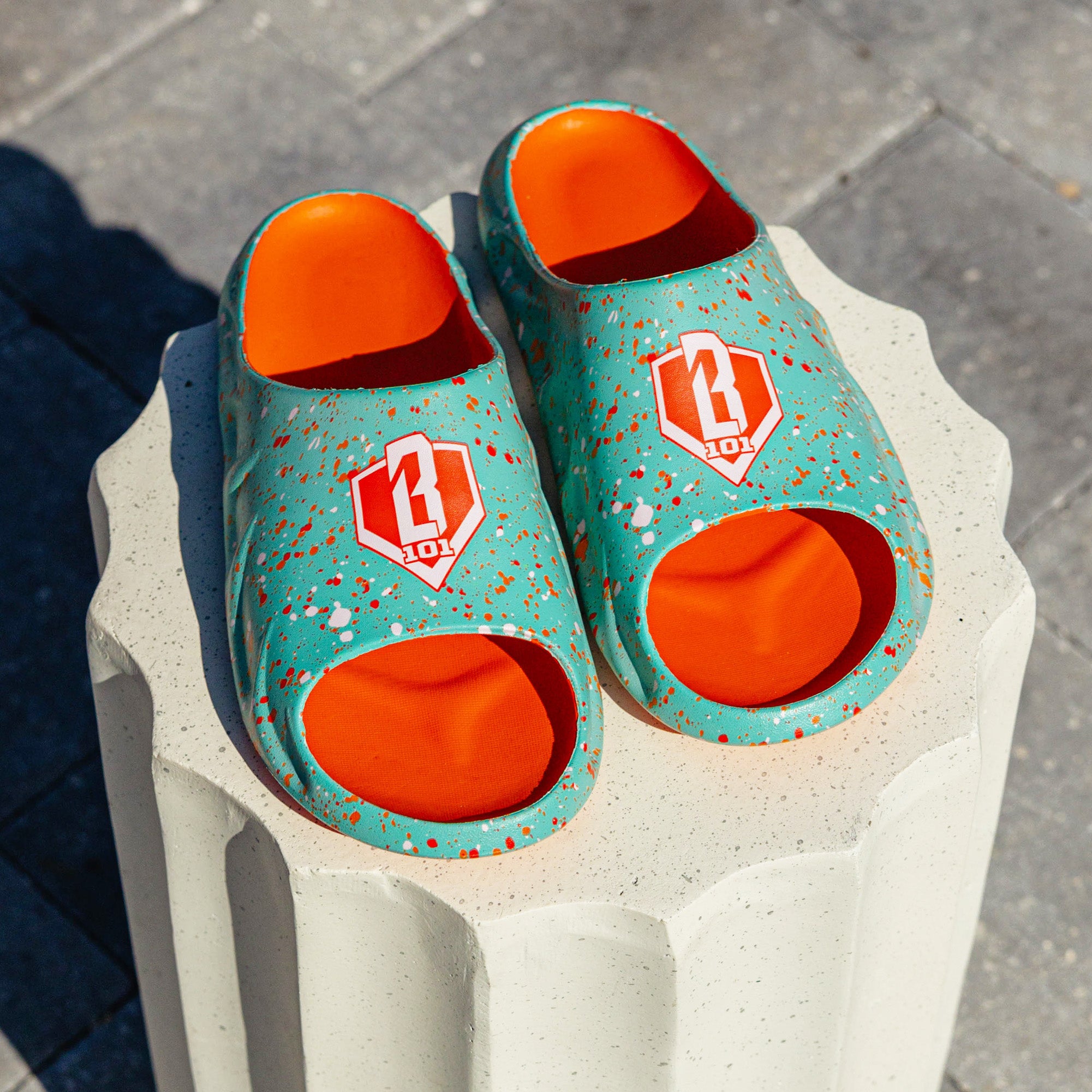 Pair of BL101 teal splatter print sandals with orange footbeds and BL101 logo, displayed on a white concrete pedestal.