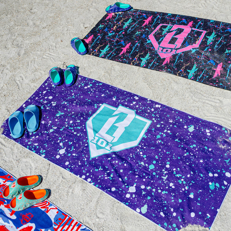 Three BL101 beach towels laid out on sand in neon shark, purple splatter, and graffiti prints, styled with colorful BL101 slides and sandals.