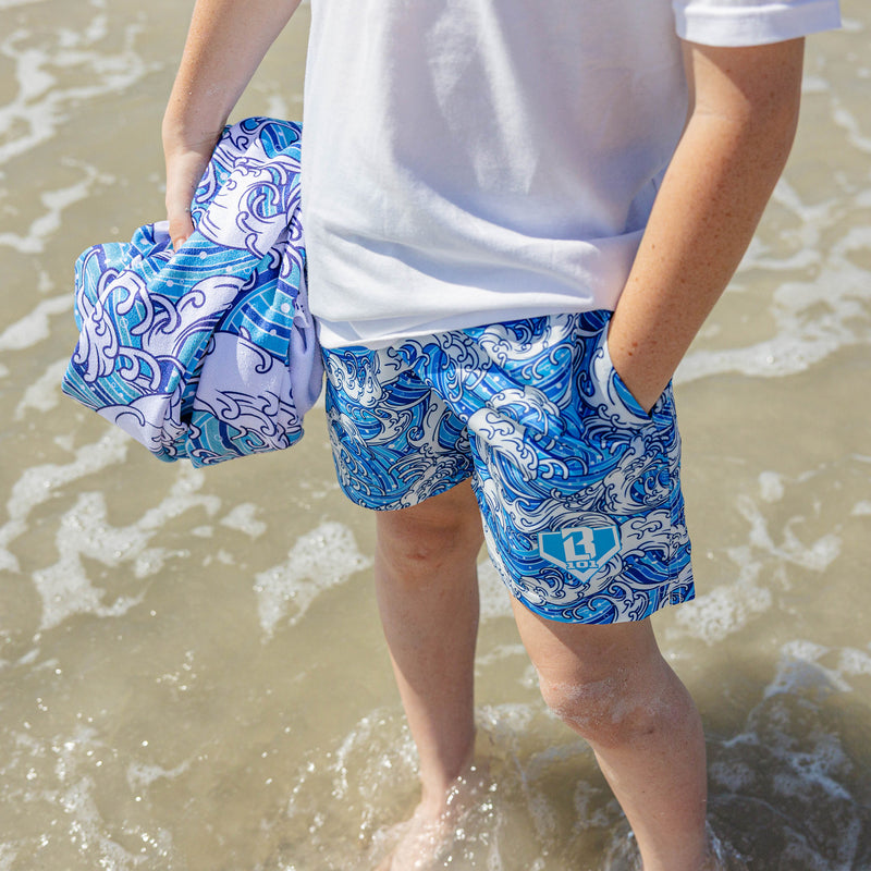 Person standing in ocean surf wearing BL101 blue wave-print swim trunks, holding a matching beach towel.
