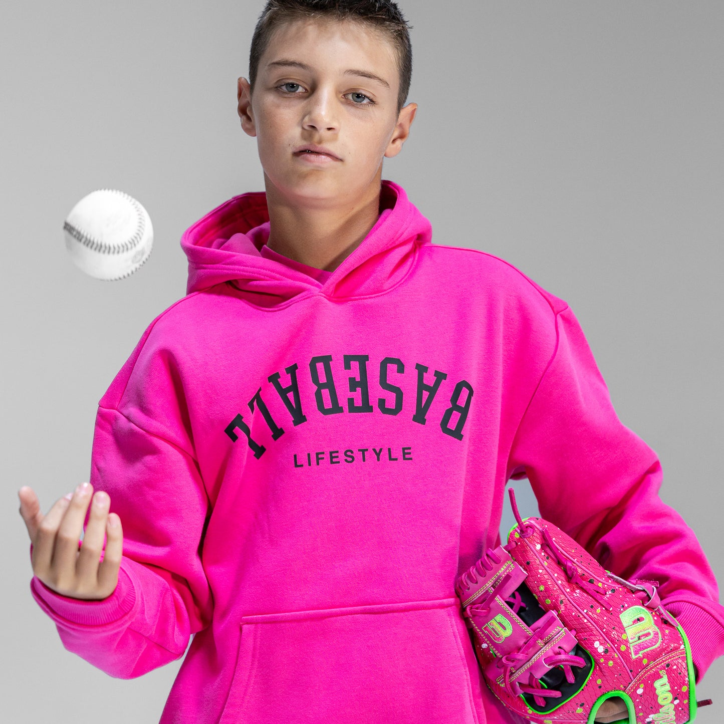 Person wearing a bright pink hoodie with upside down 'BASEBALL LIFESTYLE' text, holding a pink baseball glove against a gray background.