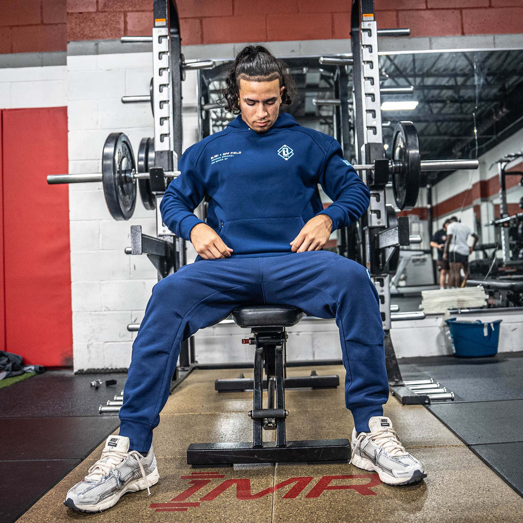 Person in blue BL101 sweatsuit, sitting on a weight bench in a gym setting
