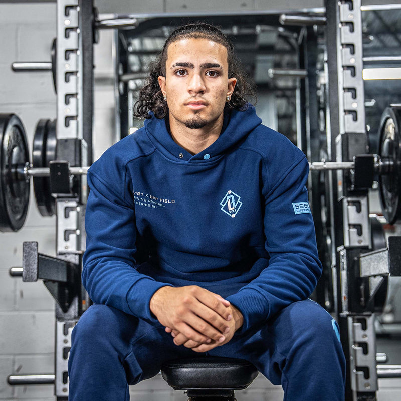 Person wearing a blue hoodie with BL101 X OFF FIELD logos sitting on a weight bench in a gym.