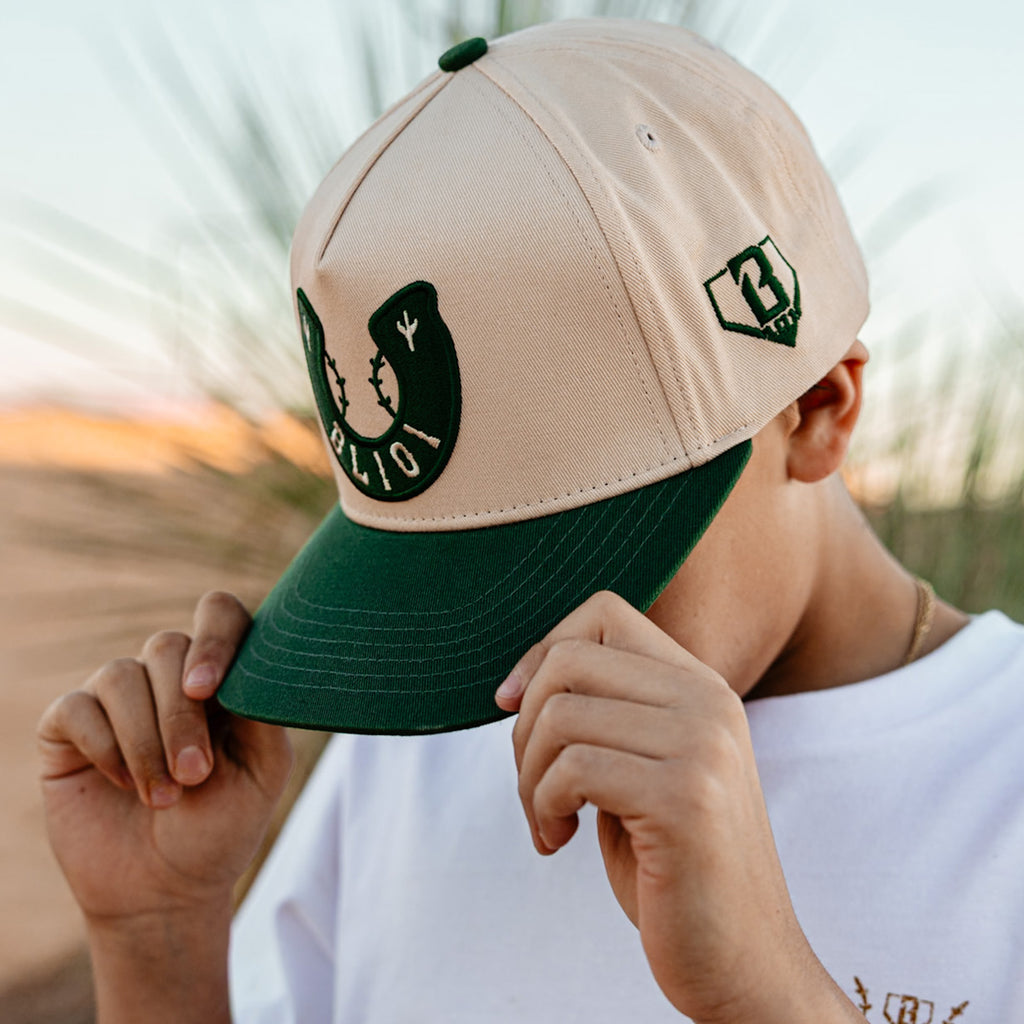 Person wearing a beige and green cap with horseshoe design and BL101 logo, holding it with both hands.