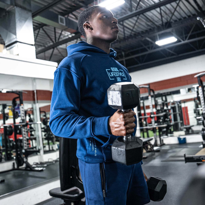 Person in a gym wearing a blue BSBL Lifestyle 101 hoodie and holding a dumbbell.