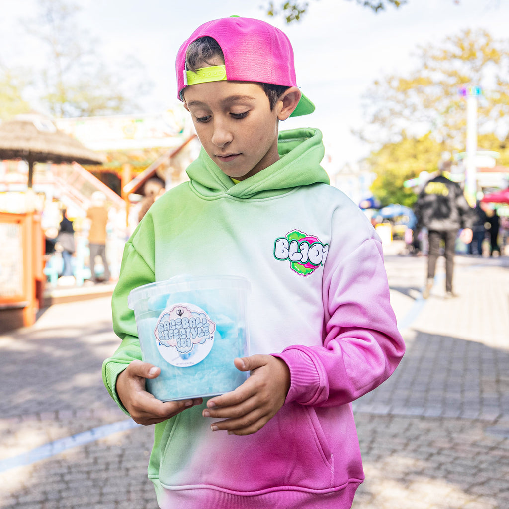 Child wearing a gradient green and pink hoodie and pink cap holding a container of cotton candy.