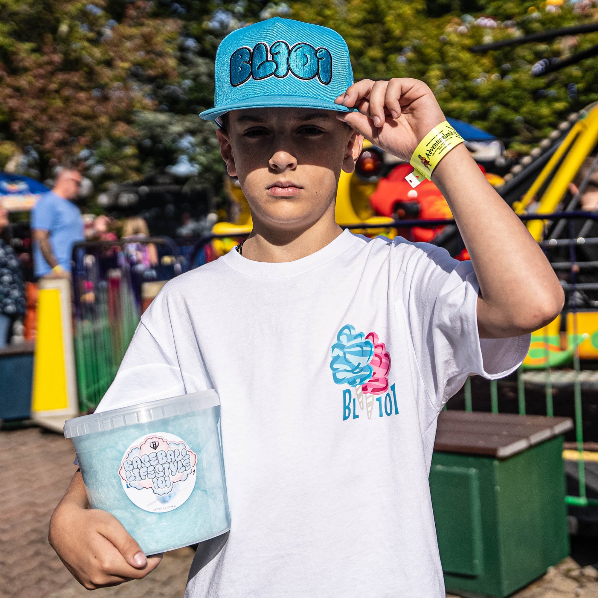 Child wearing a blue cap and white t-shirt with branding, holding a container, in an outdoor setting.