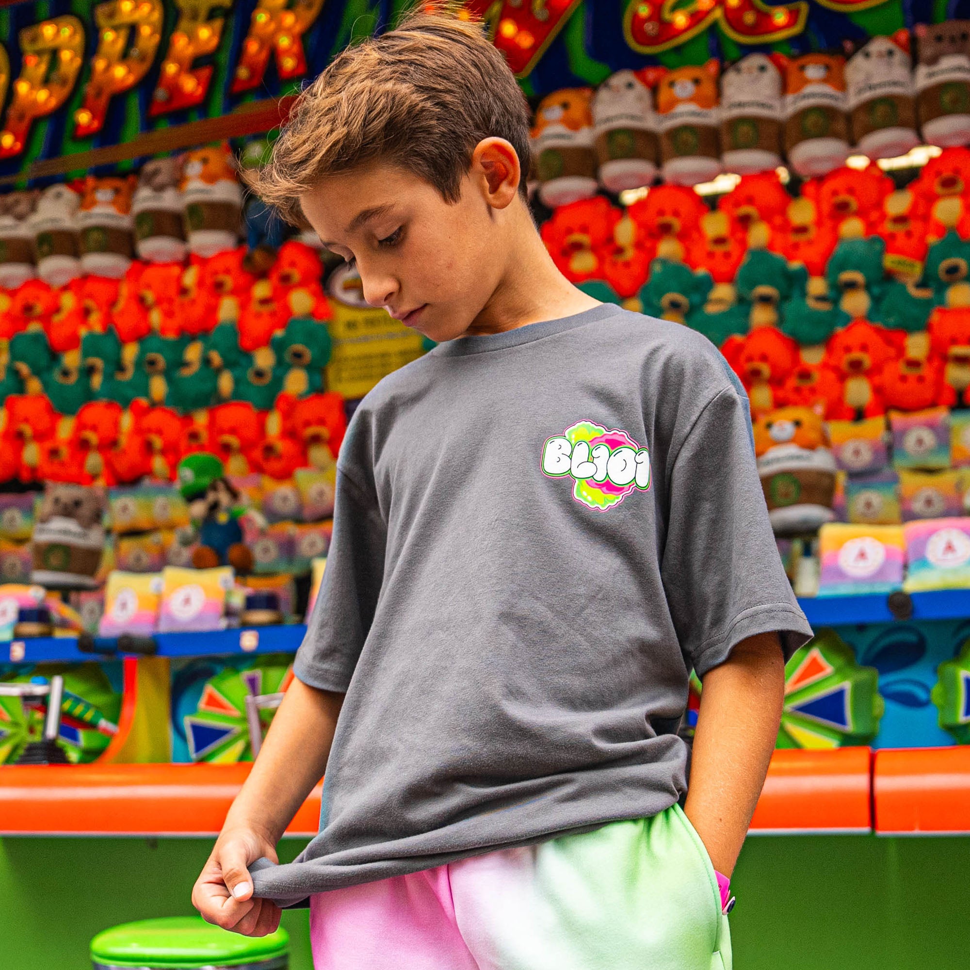 Child wearing a gray t-shirt with a pink and green BL101 logo in front of a colorful toy display.