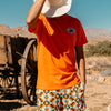Person wearing an orange t-shirt with a 'WHERE DUST MEETS DIAMOND' 
logo and patterned shorts in a desert setting with a wooden cart and mountains.