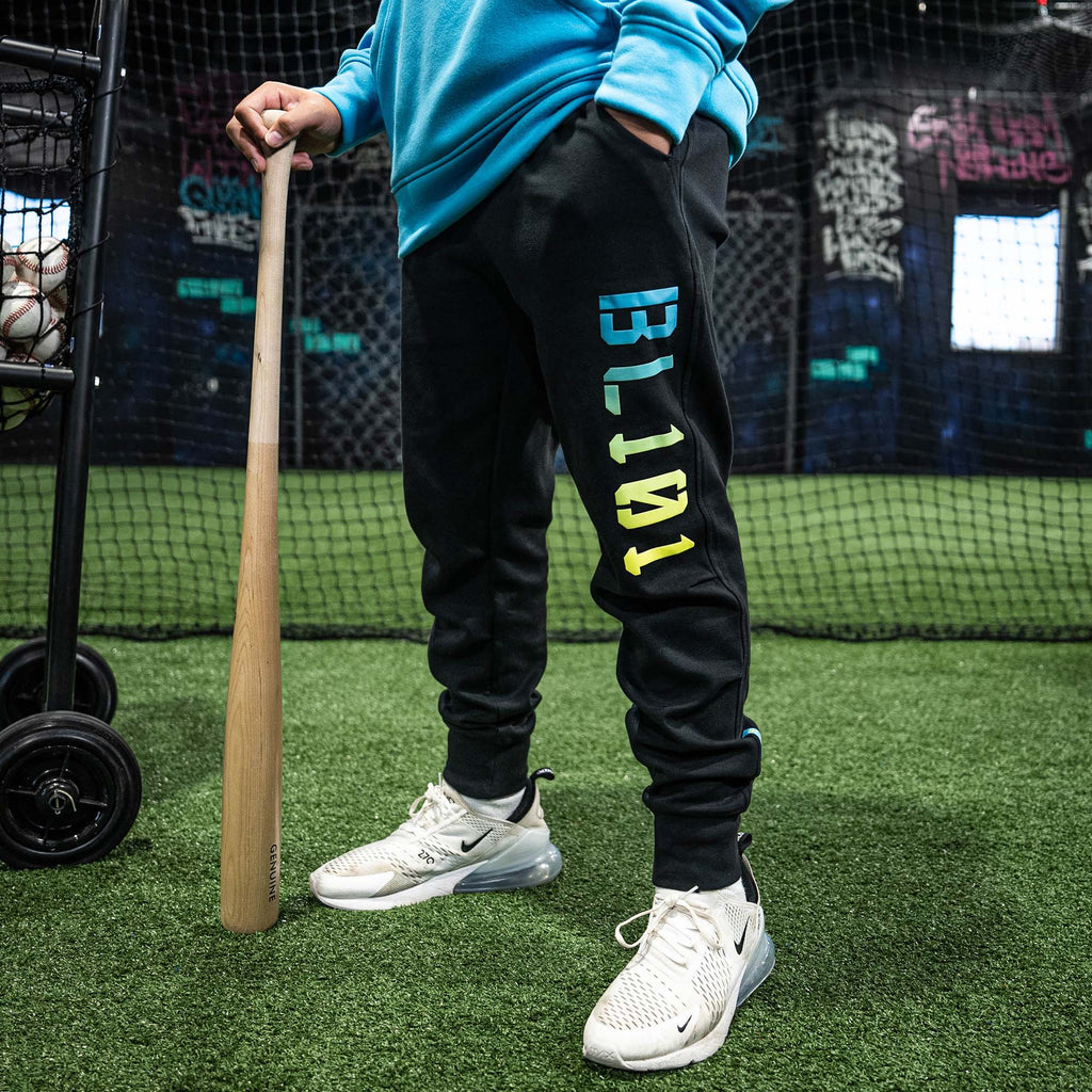 Person wearing black pants with colorful 'BL101' text on leg, holding a baseball bat in an indoor sports facility.
