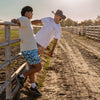 Two people wearing blue and orange patterned shorts on a dirt path with a wooden fence and open field in the background