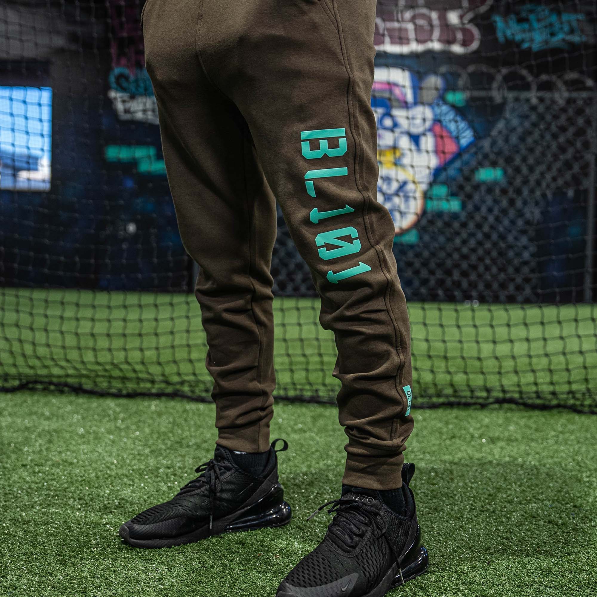 Person wearing brown jogger pants with 'BL101' branding on grass, with a colorful background.