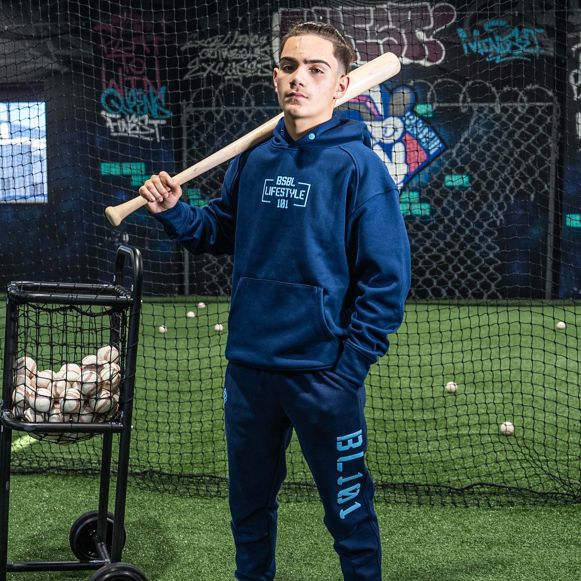 Person wearing a blue hoodie and sweatpants with BL101 logo holding a baseball bat in an indoor batting cage.