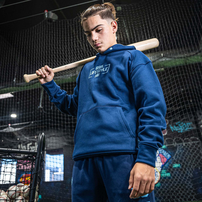 Person wearing a blue hoodie with a lBSBL Lifestyle 101 logo, holding a baseball bat in an indoor sports facility.