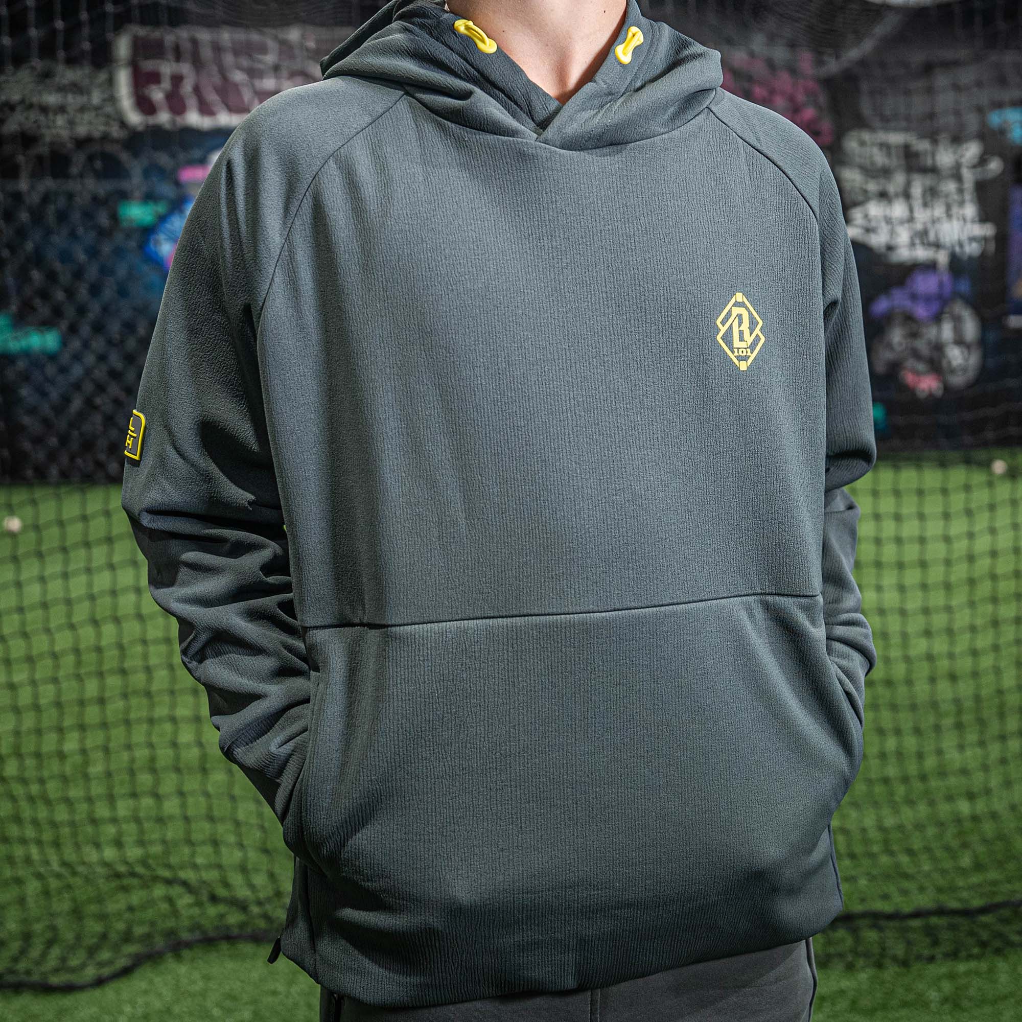 Person wearing a gray hoodie with a yellow BL101 logo in front of a sports net.