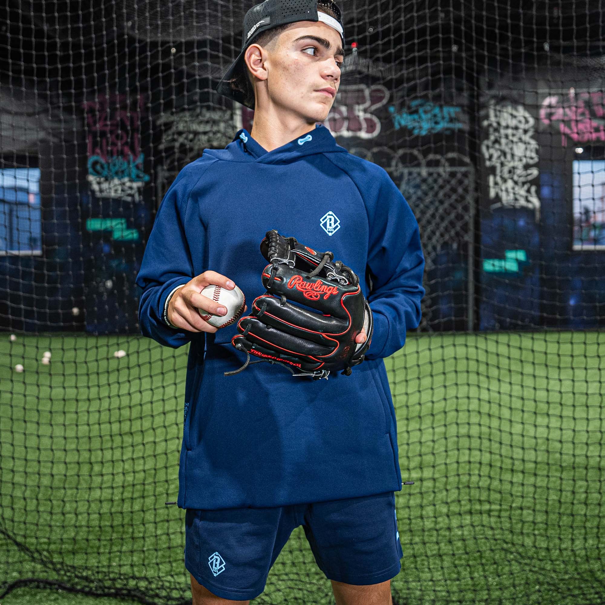 Person in blue BL101 athletic wear holding a baseball and glove in front of a batting cage.