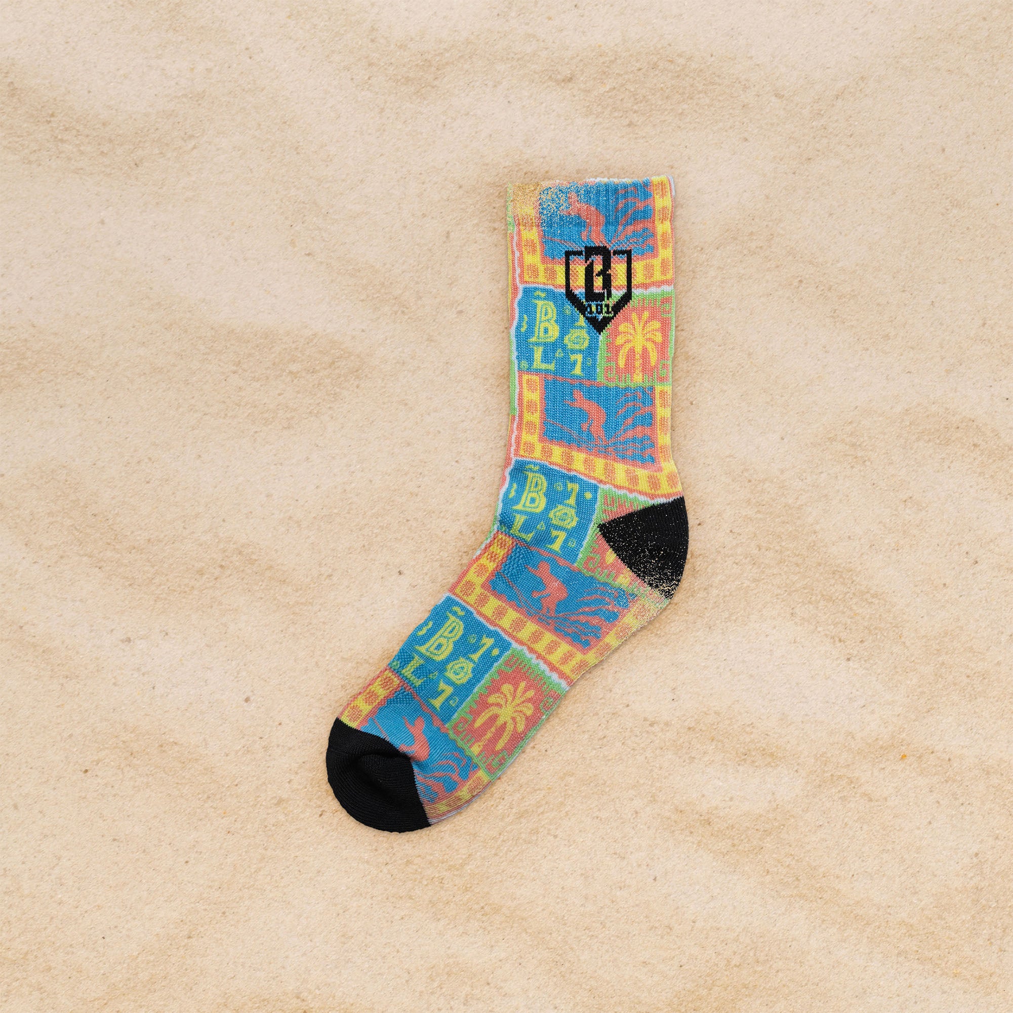 A single crew sock with a colorful patchwork design featuring blue, yellow, orange, and green blocks with palm tree and sun motifs, finished with black toe and heel caps, laid flat on a sandy beige background.
