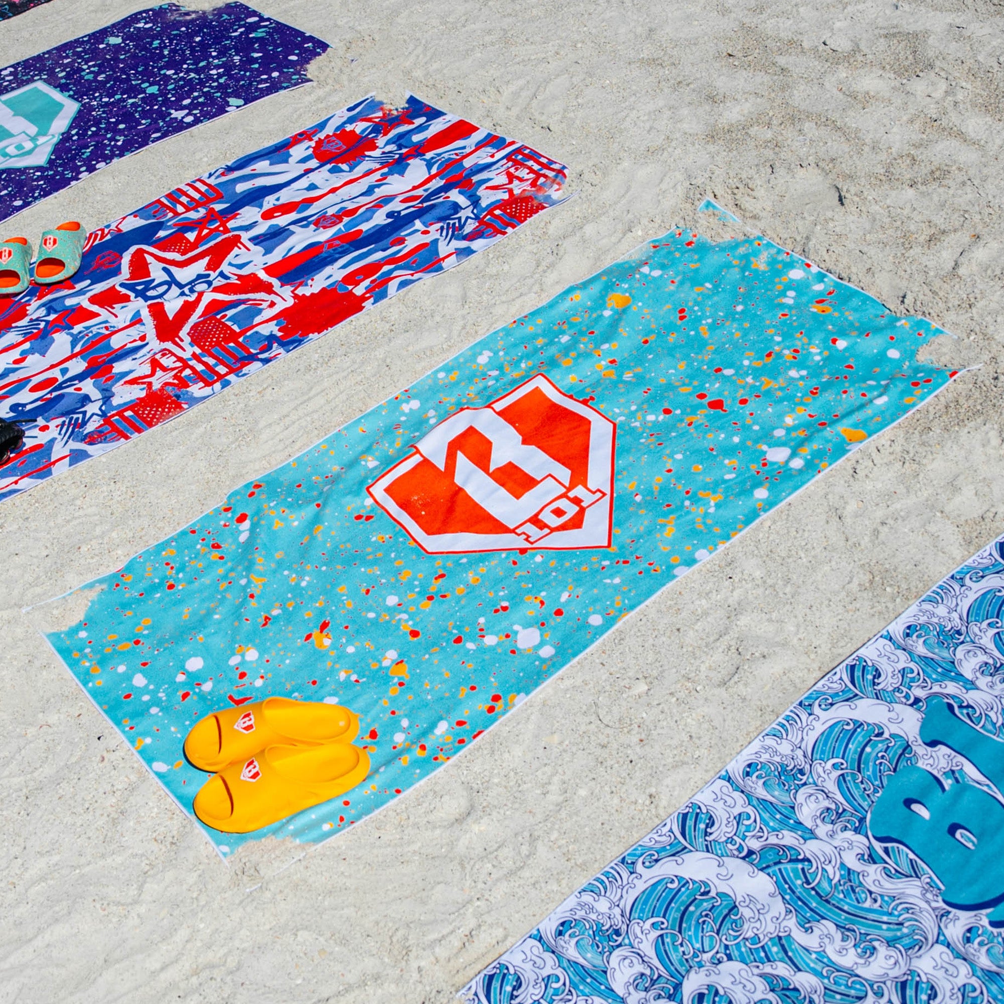 Three BL101 beach towels laid out on sand in graffiti, teal splatter, and wave prints, styled with yellow and green BL101 slides.