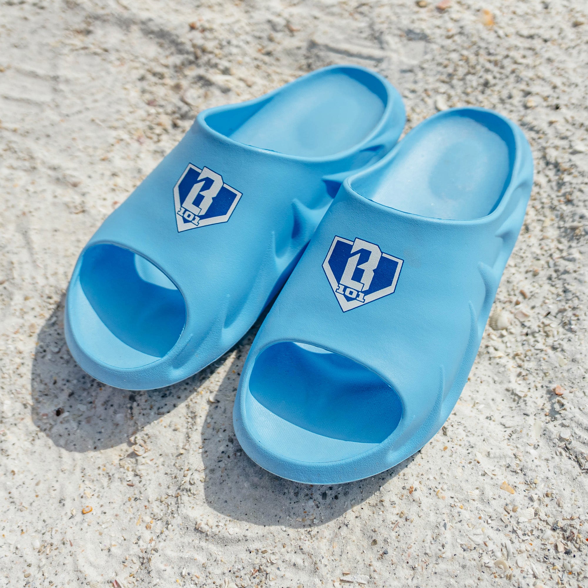 Pair of BL101 light blue slides on sand with the blue and white home plate logo on each strap.