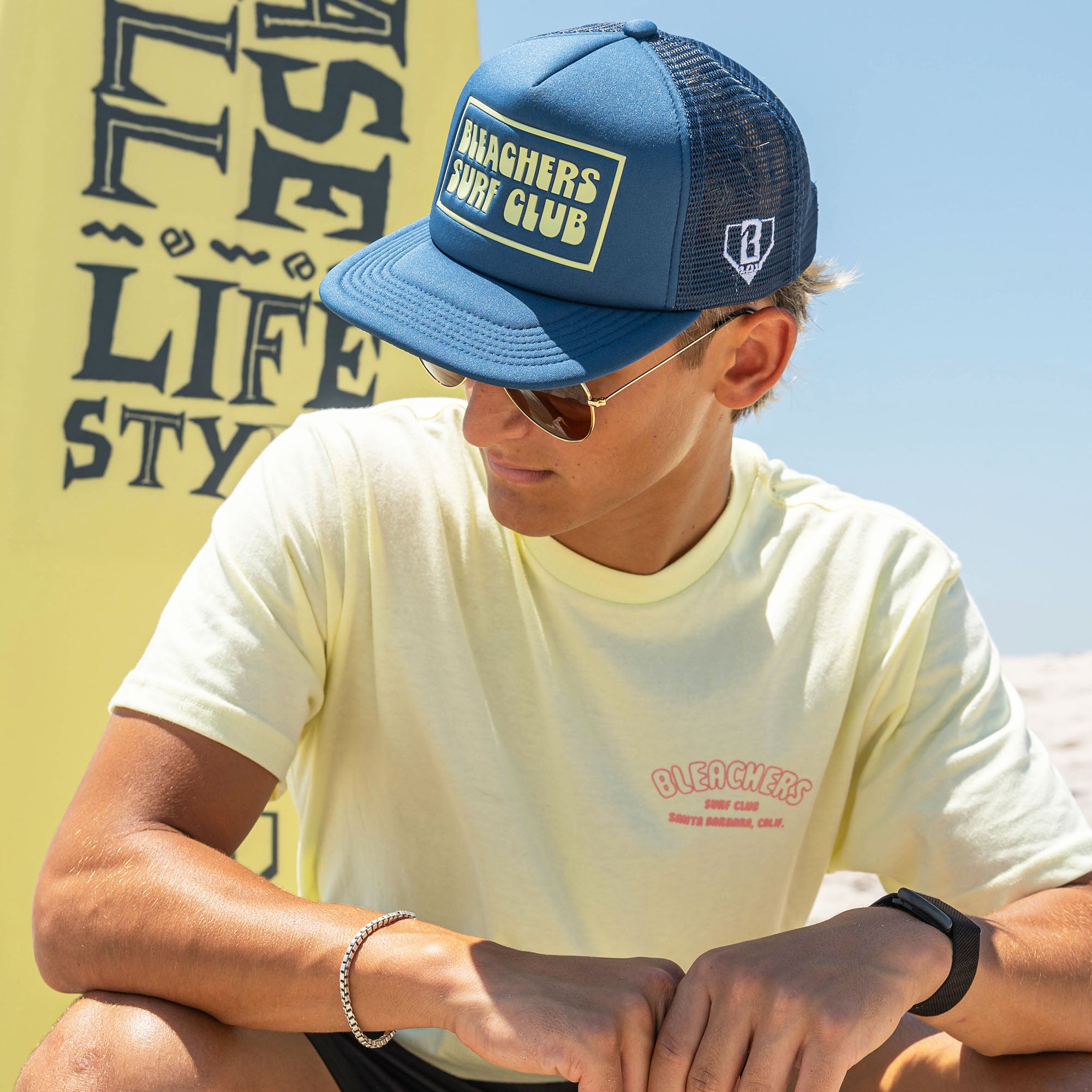 Adult model wearing blue hat with "Bleachers Surf Club" design