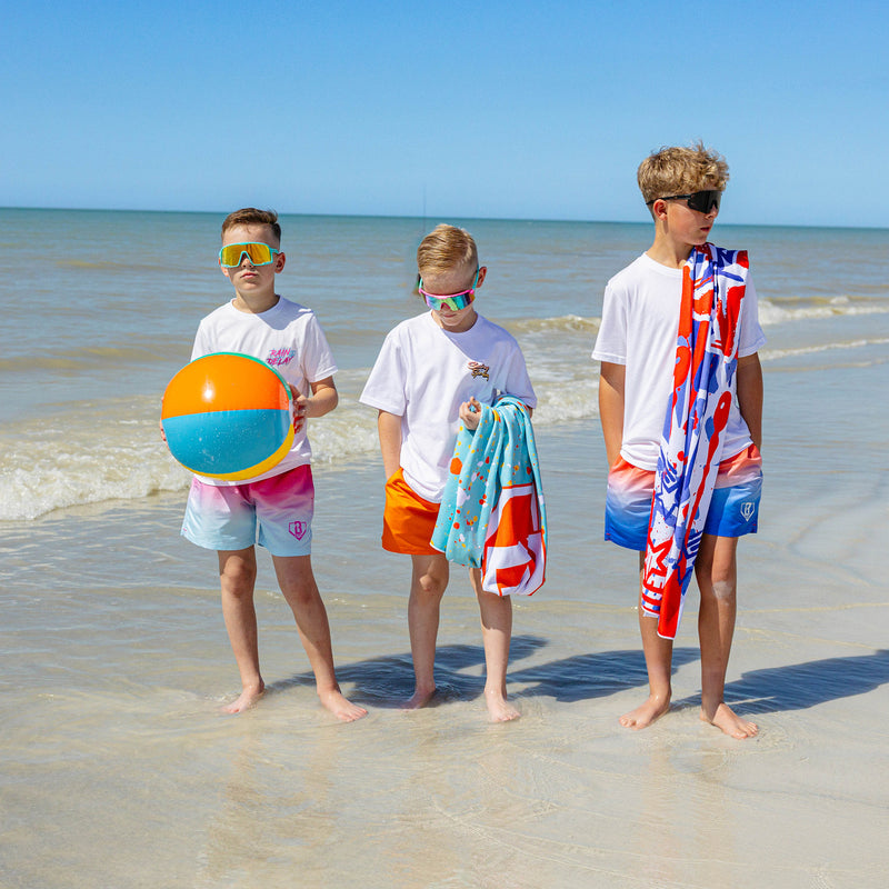 Three boys standing at the shoreline in BL101 swim trunks and graphic tees, wearing sunglasses, holding a beach ball, a splatter-print towel, and a graffiti-print towel.
