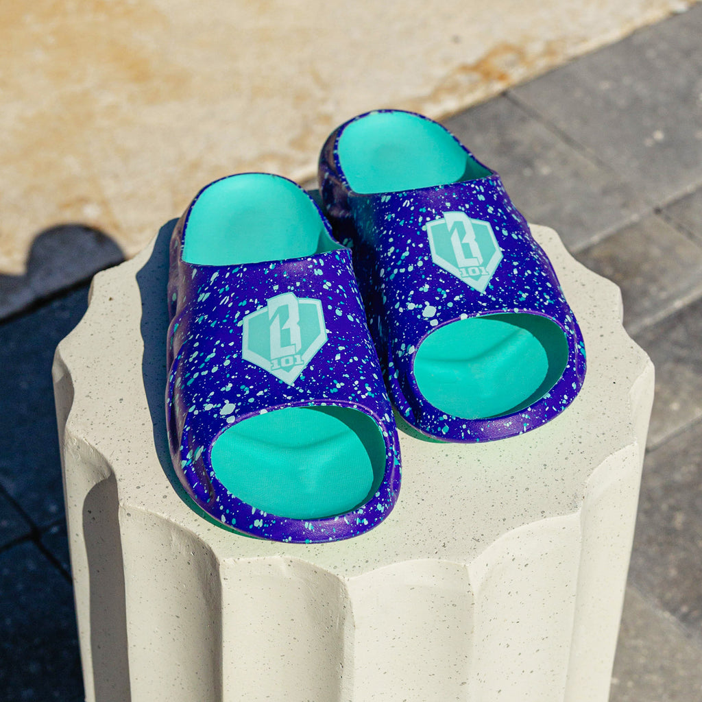 Pair of BL101 purple splatter slides with teal footbeds displayed on a white concrete pedestal, shot from the front.