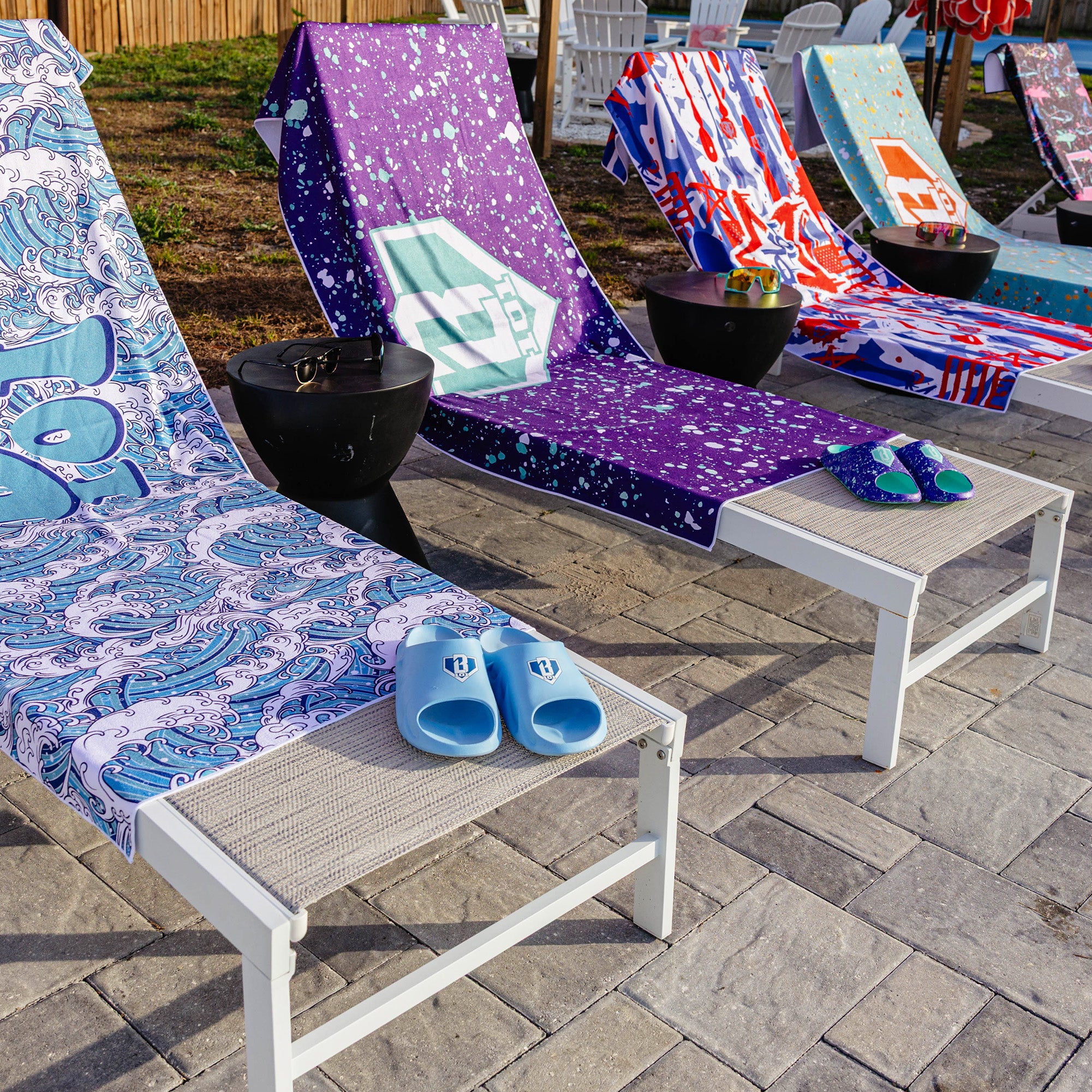 Four BL101 beach towels draped over white outdoor lounge chairs in wave, purple splatter, graffiti, and teal splatter prints, with BL101 slides on the chairs.