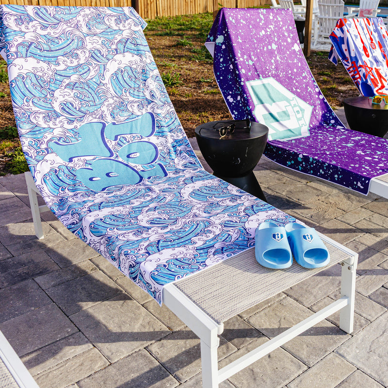 Three BL101 beach towels on outdoor lounge chairs — wave print, purple splatter, and red/white/blue — with light blue BL101 slides in the foreground
