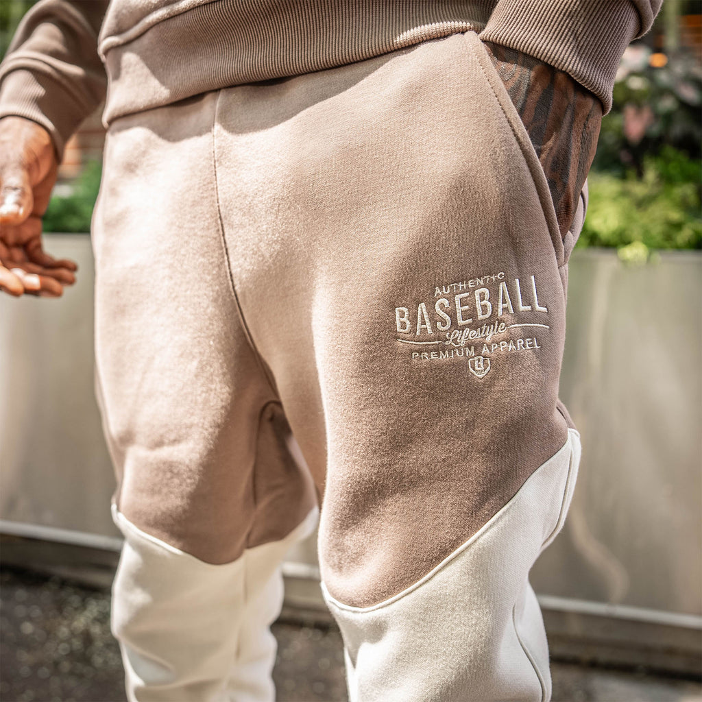 Beige sweatpants with 'Baseball Lifestyle PREMIUM APPAREL' logo worn by a person outdoors.