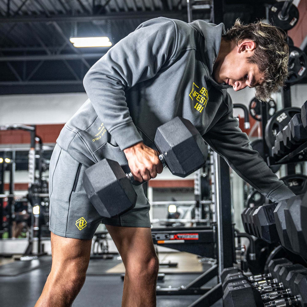 Man lifting dumbbells wearing a gray BL101 hood with yellow logo in a gym setting