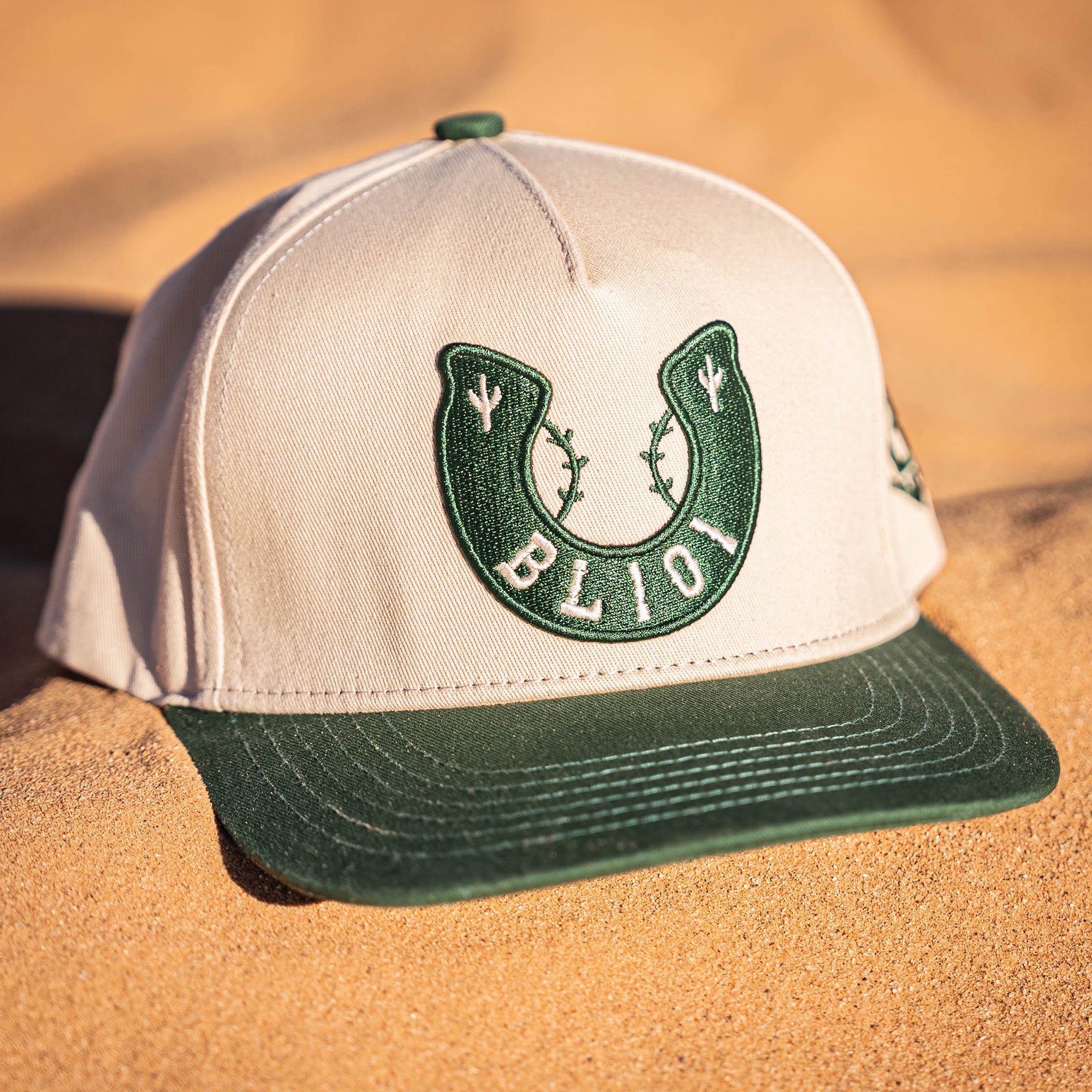 Beige and green cap with a horseshoe design and BL101 logo on a sandy surface