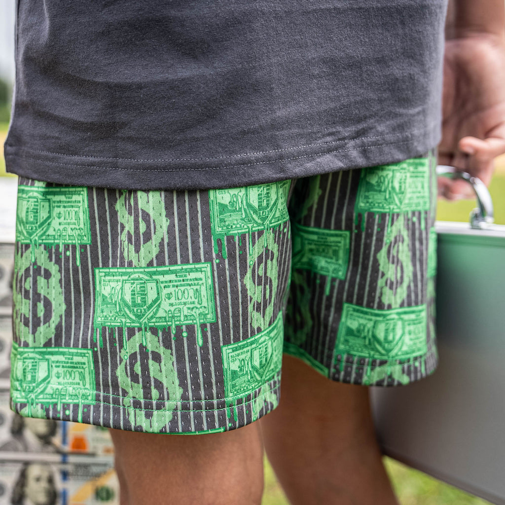 Person wearing green and gray patterned shorts with dollar bills on it, holding a suitcase