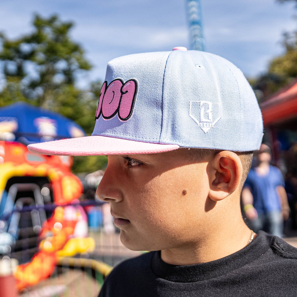 Person wearing a light blue cap with pink text and a logo, standing in an outdoor setting with trees and a colorful structure in the background.