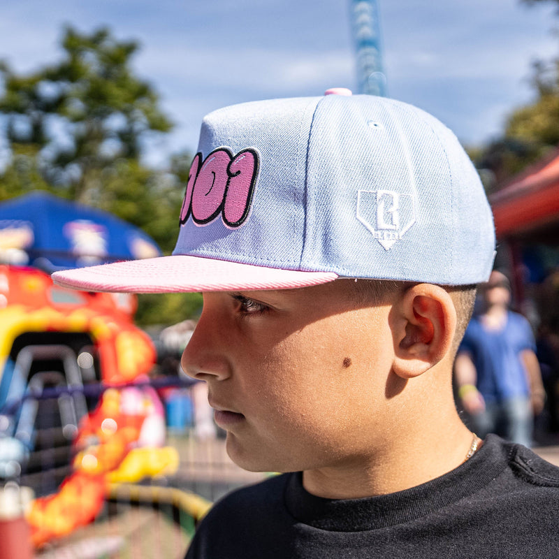 Person wearing a light blue cap with pink text and a logo, standing in an outdoor setting with trees and a colorful structure in the background.