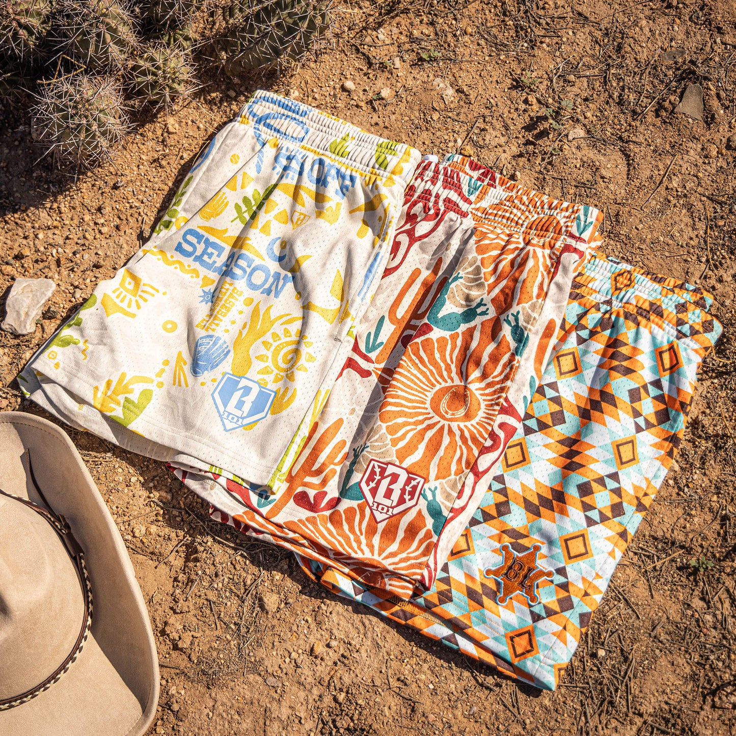 Three pairs of patterned shorts on a sandy surface with a hat nearby.