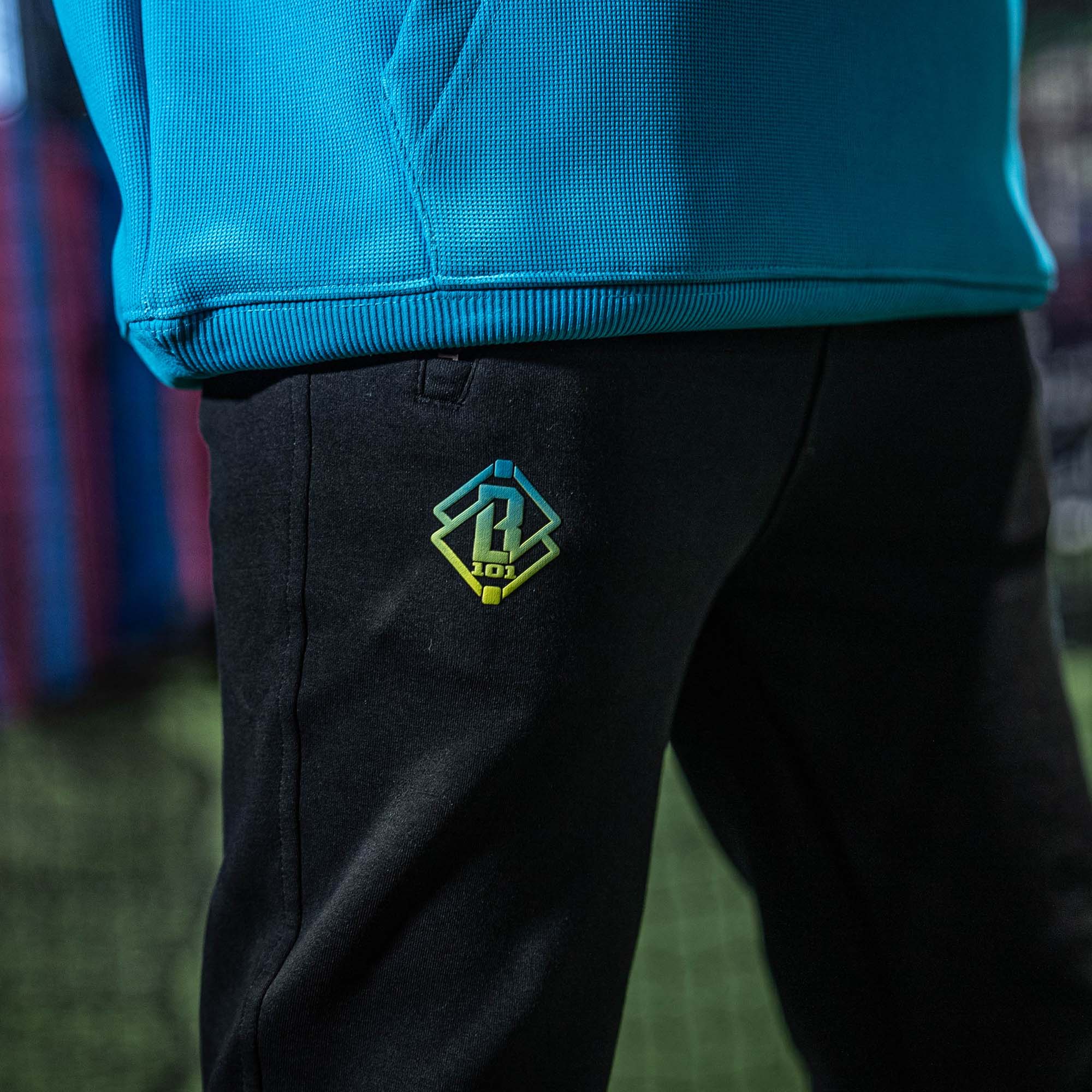 Black pants with a green and blue BL101 logo , worn with a blue jacket, against a blurred background.