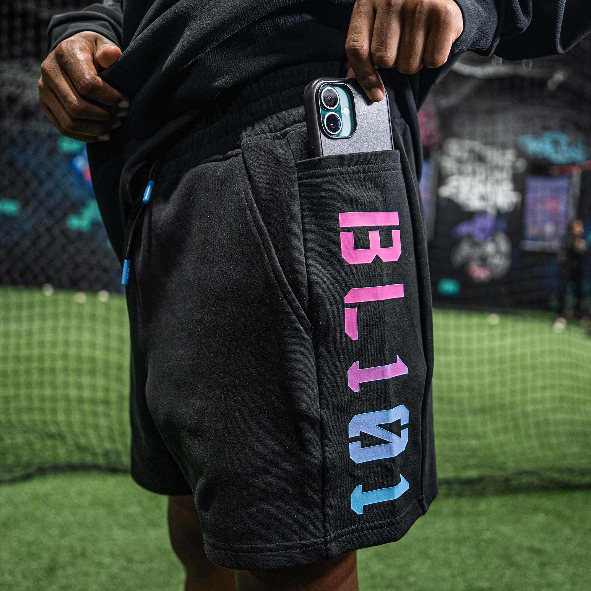 Person wearing black shorts with colorful BL101 text, holding a phone, on a sports field.