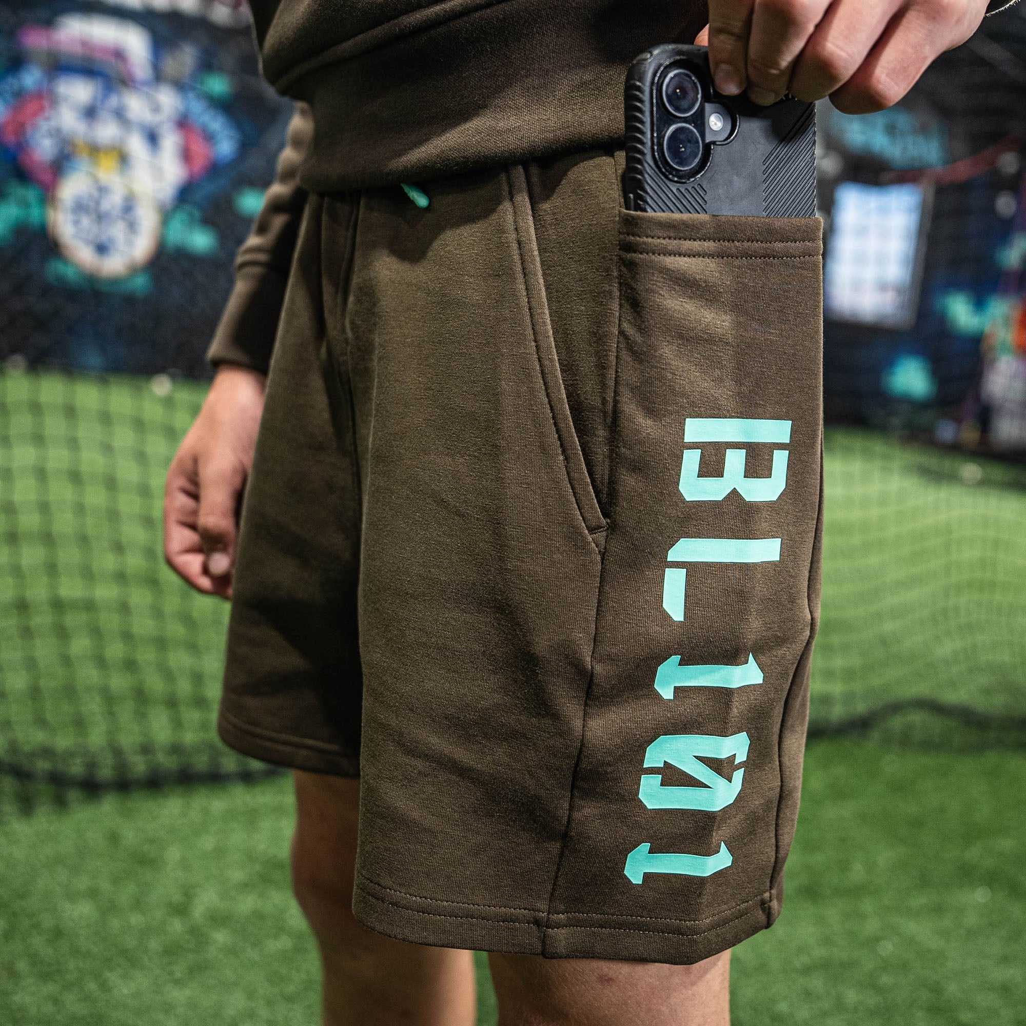 Person wearing brown shorts with 'BL101' branding, holding a phone, on a grassy field.